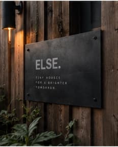 Schild aus dunklem Metall mit „ELSE.“ an einer Holzwand, daneben eine Lampe